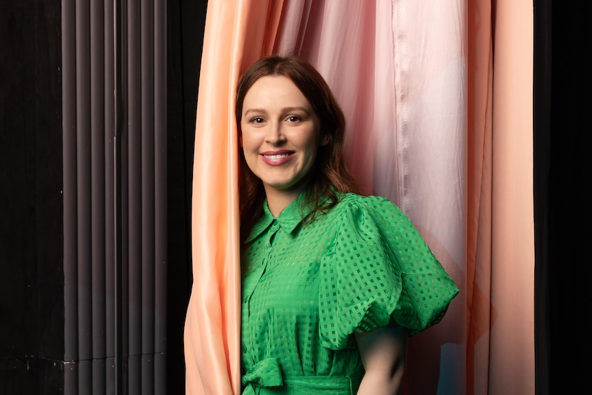 Laura Murphy, a brunette white woman in her 30s, wears a bright green dress and stands between pink stage curtains.