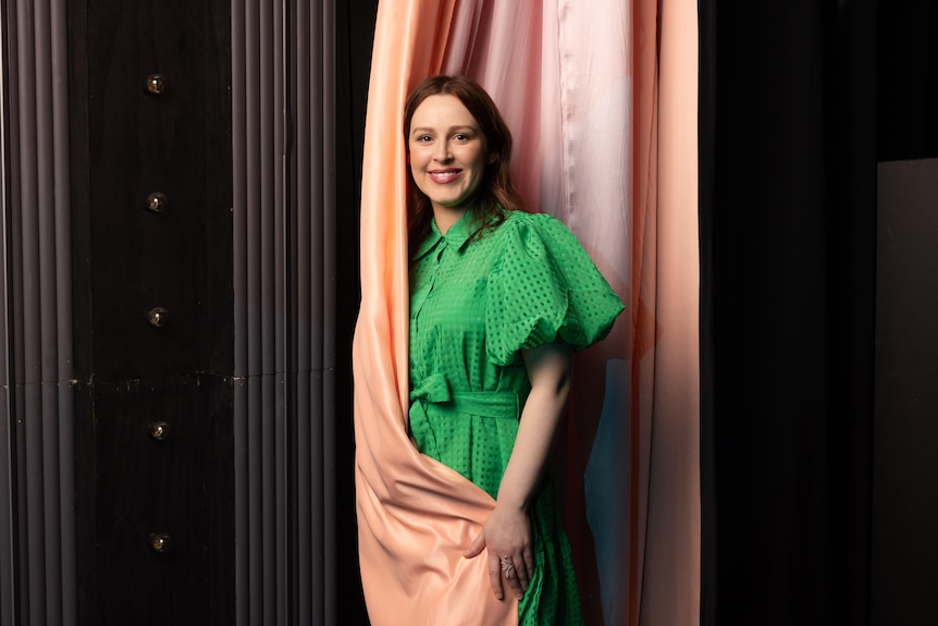 Laura Murphy, a brunette white woman in her 30s, wears a bright green dress and stands between pink stage curtains.