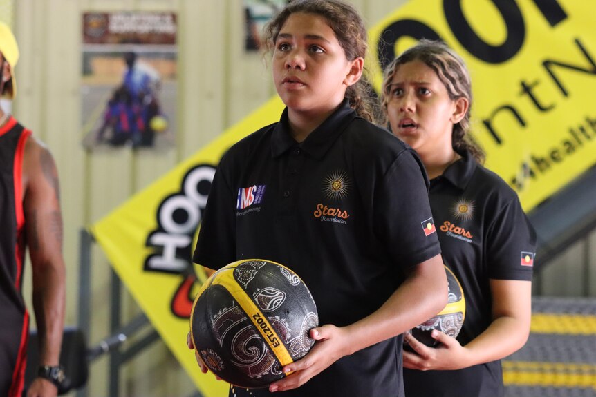 A young Indigenous girl holding a basketball in a gymnasium, looking up at a hoop and ready to shoot.