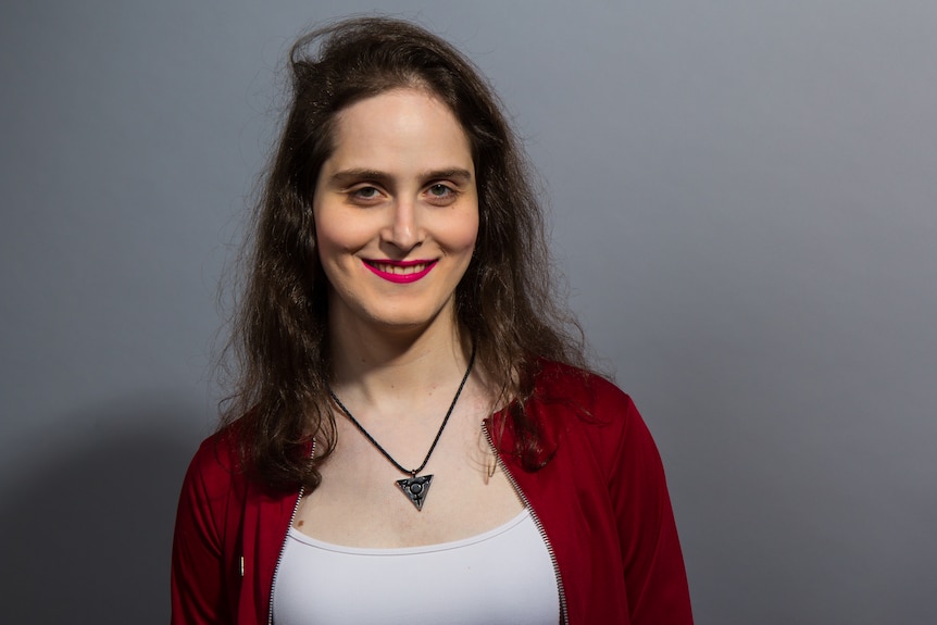 A young woman with long dark hair and red lipstick smiles as she tilts her head.