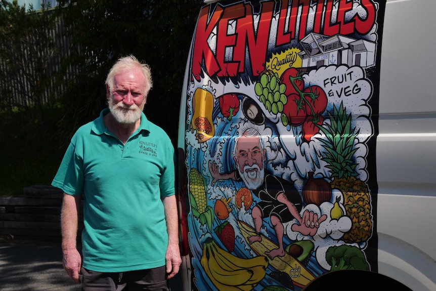 A man in his sixties stands next to a delivery van, with a painted sign on it for a fruit and vegetable shop.