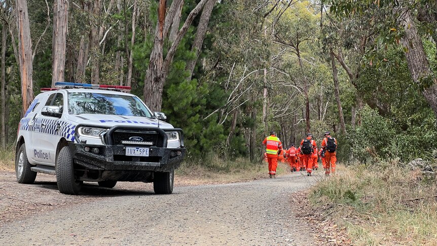 A police vehicle blocks a dirt road with SES volunteers wearing orange in the background.
