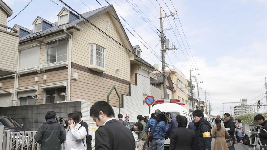People and reporters standing outside a weatherboard apartment in Japan.