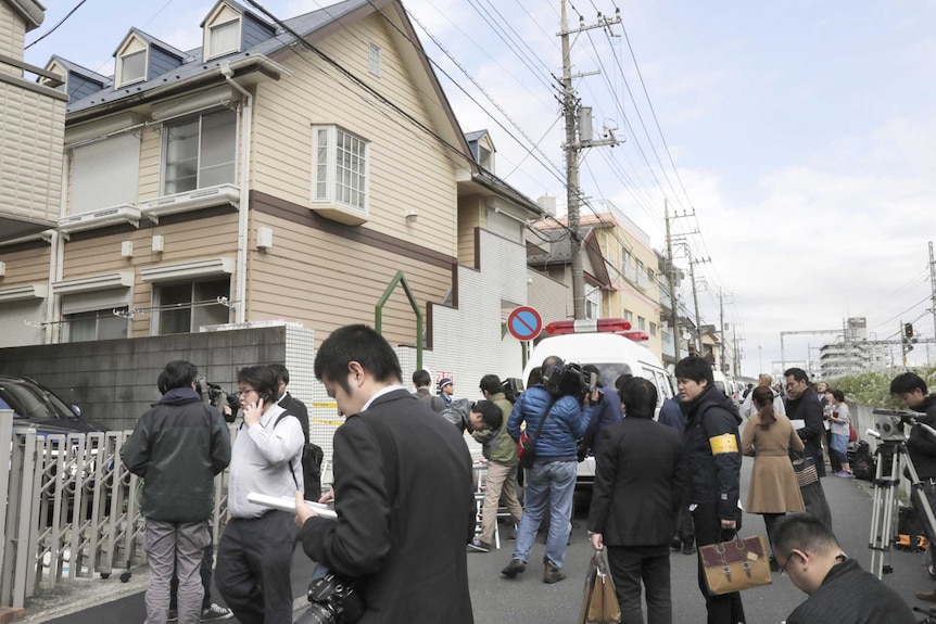 People and reporters standing outside a weatherboard apartment in Japan.