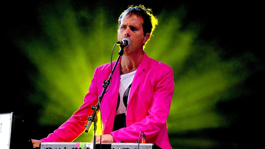 Julian Hamilton of The Presets wears a pink jacket, singing and playing keys as green light explodes behind him