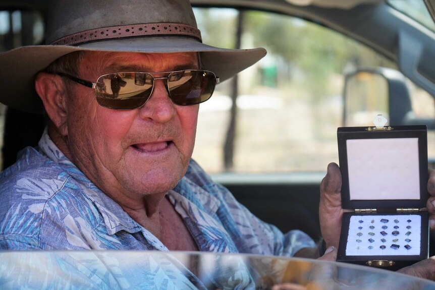 Older man wearing hat, sunglasses, holds jewellery box of precious cut gems while sitting in a car.