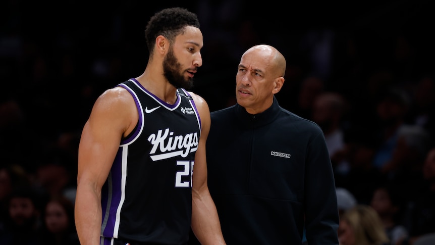 Two men (one very tall and wearing basketball team kit) speak to each other sharing frustrated facial expressions.