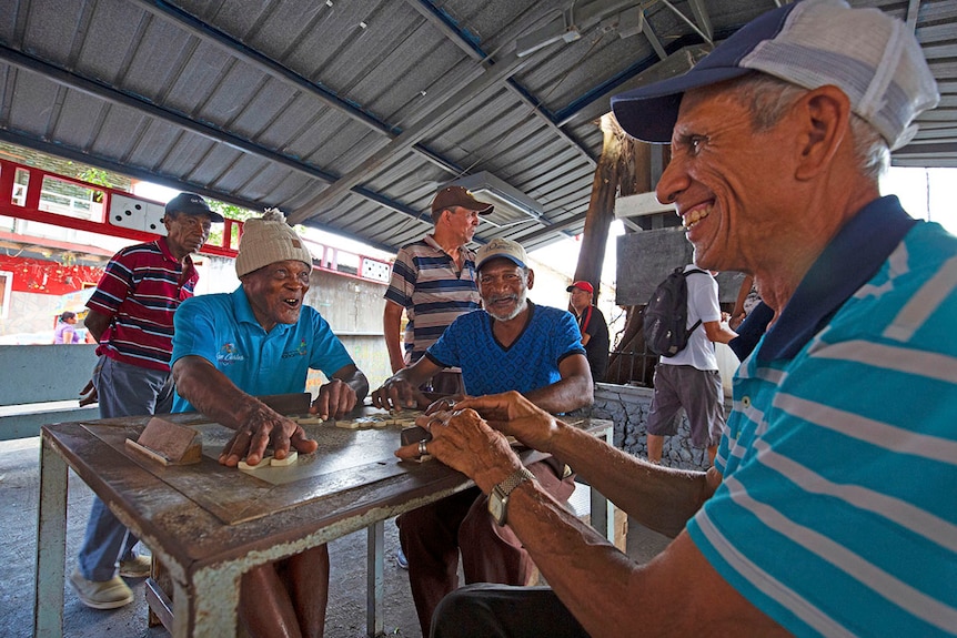 Three men smile around a domino board.