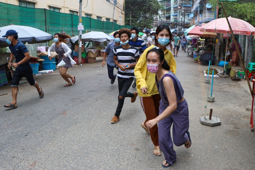 Young people in face masks run in a street with market umbrellas in the background. 