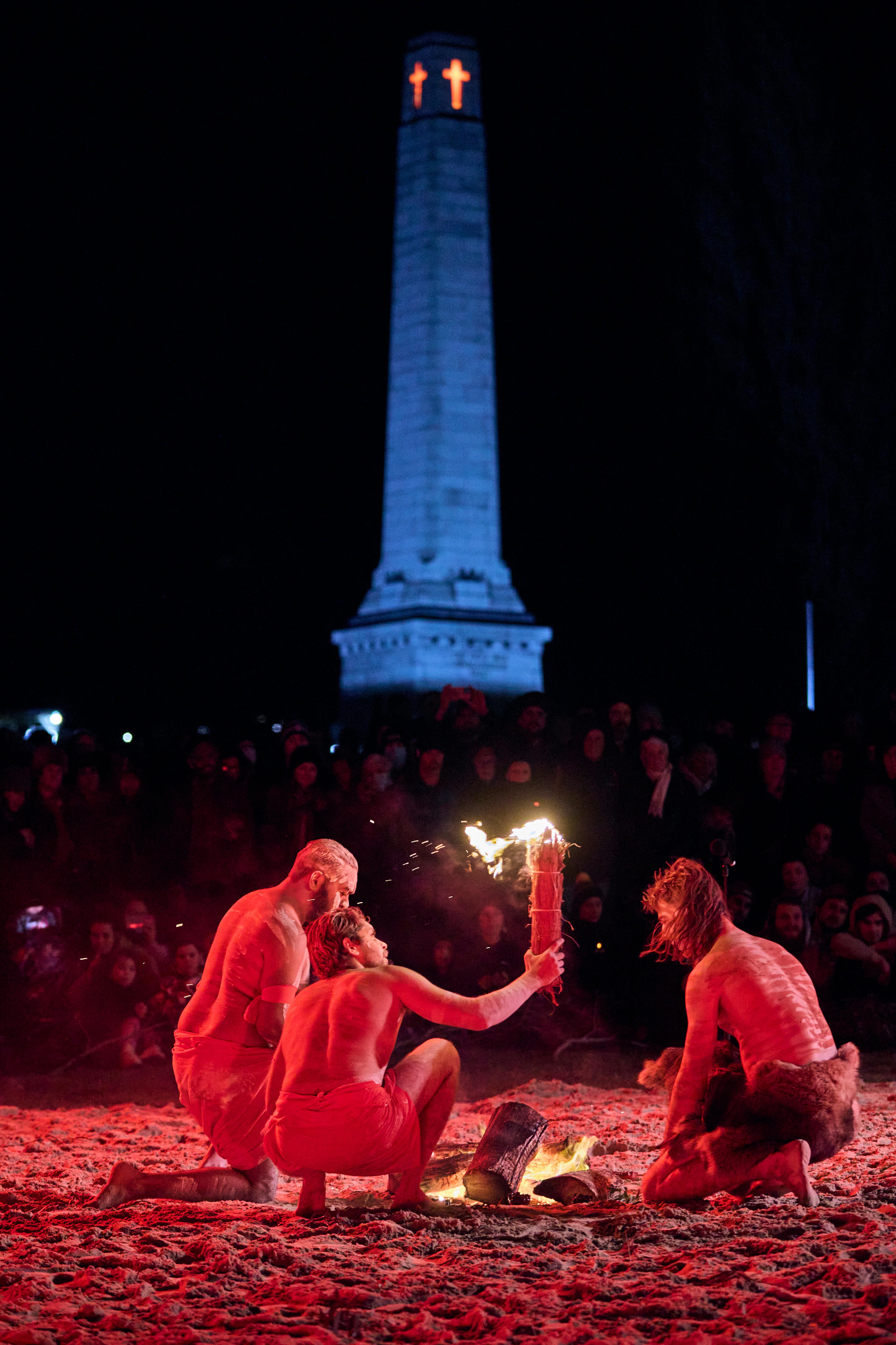 Three First Nations performers crouch in soil around a fire before an audience. One holds a flaming torch.