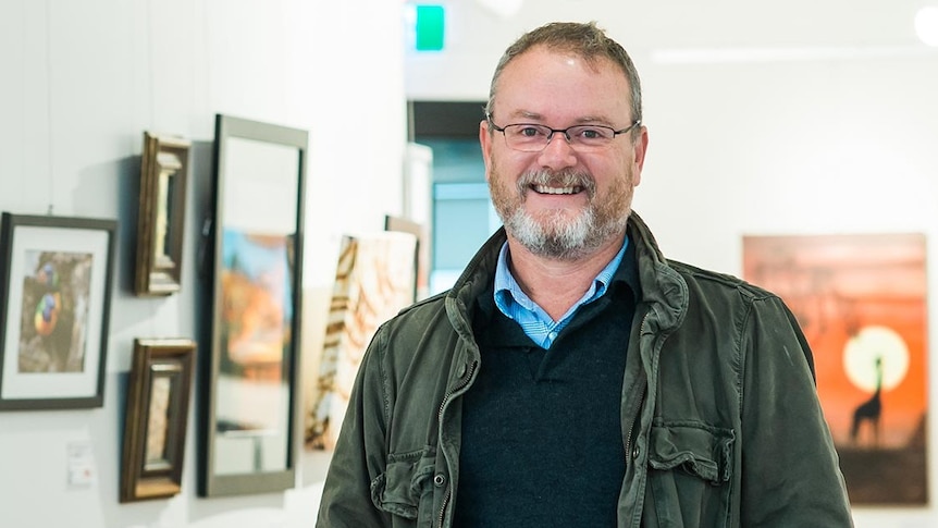 A man in a knit sweater, khaki jacket and glasses smiles in a brightly lit gallery with framed imaged behind him.