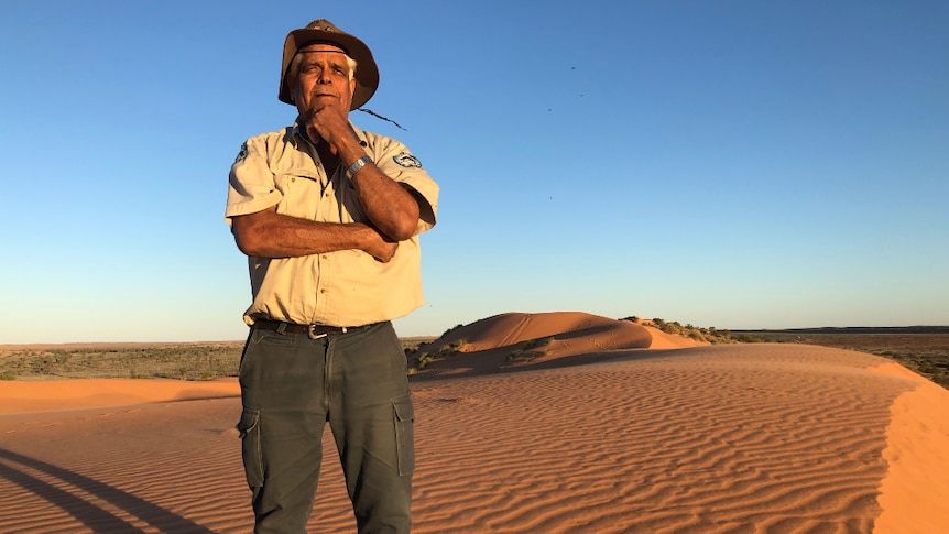 A ranger stands on a sand dune in the desert, looking into the distance with his hand on his chin.