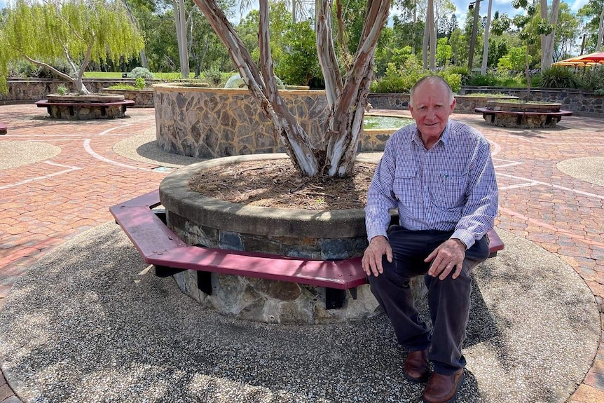 A man sits in a paved area with trees.