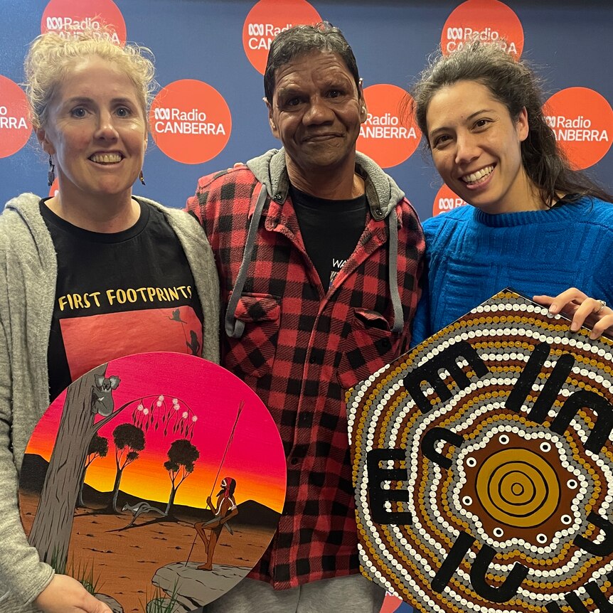 Two women and a man stand against ABC Radio Canberra's backdrop and hold two artworks created by Wahluba artist William Walker