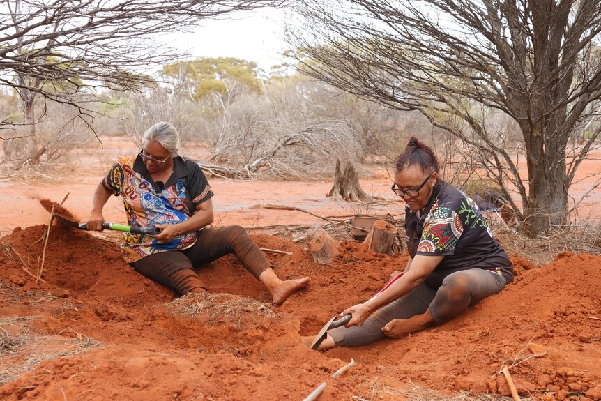 two women digging in red dirt under trees 