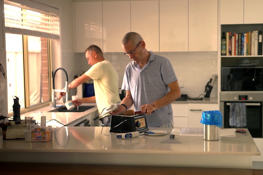 A man fills a cup with water from a kitchen tap while another man prepares toast.