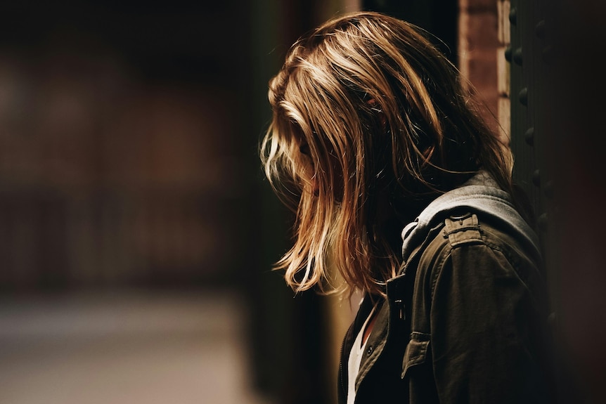 A teen girl with hair over her faces looks down at the ground in a dimly lit street.