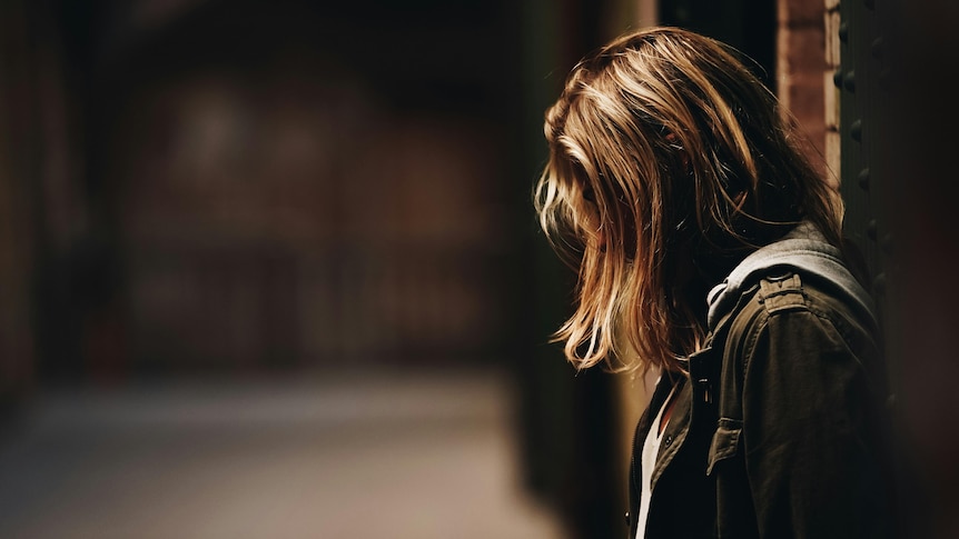 A teen girl with hair over her faces looks down at the ground in a dimly lit street.