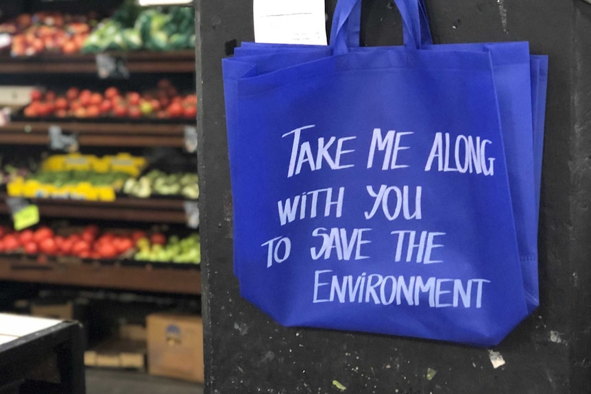 A blue reusable shopping bag which says take me along with you to save the environment hanging on the wall at a fruit shop.