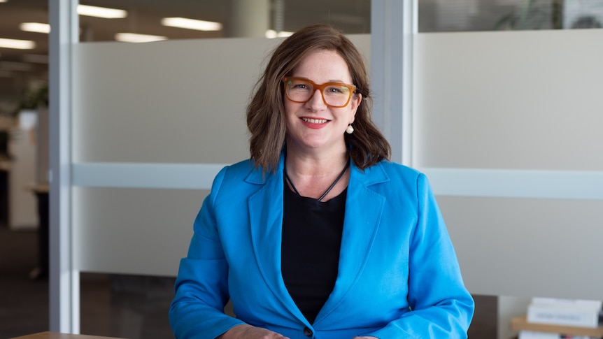 RACGP president Karen Price sits at a desk with a pen in her hand, looking into camera.