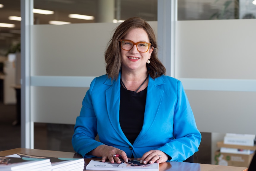 RACGP president Nicole Higgins sits at a desk with a pen in her hand, looking into camera.