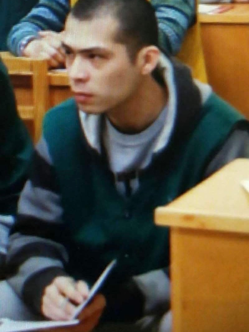 A man in jumpsuit sits near a desk holding a pen and a notebook.