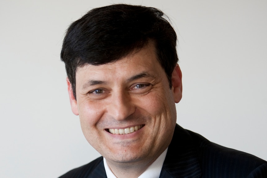 A man in a dark suit smiles as he poses for a head shot in front of a white background. 