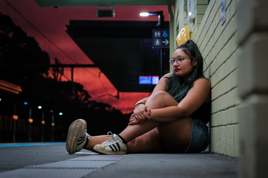 Tee sitting on the ground at a train station, leaning against a brick wall.