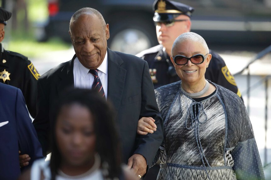 Bill Cosby smiles arm-in-arm with wife Camille as they walk. They are flanked by police officers.