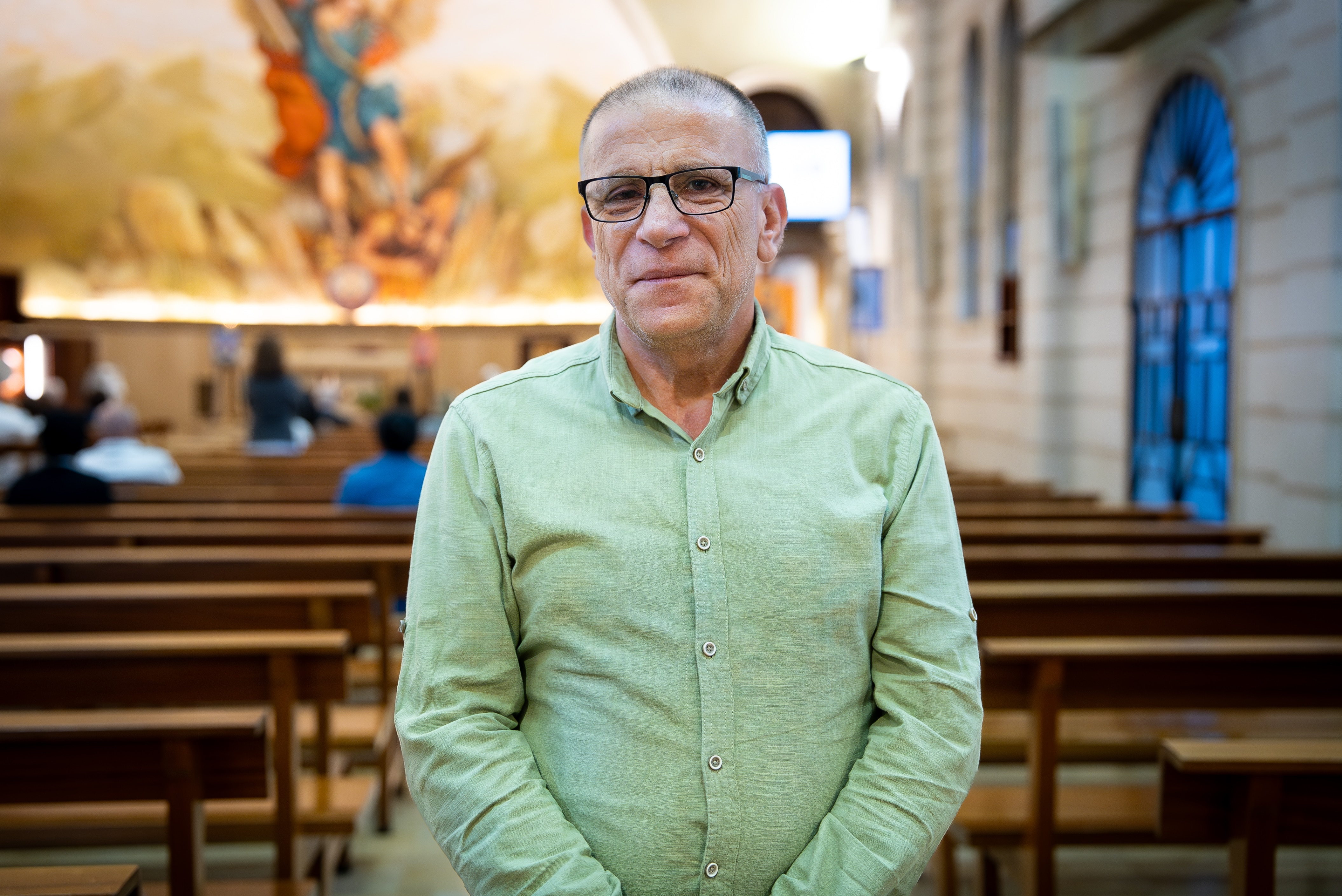 A man wearing a green collared shirt and glasses stands in a church in front of the benches