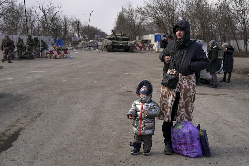 A man and a small child wearing large puffy jumpers stand on the side of a dirt road with bags. a tank can be seen behind them