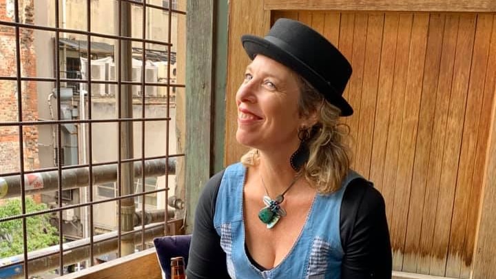 A smiling woman in a hat sitting on a bench drinking a beer.