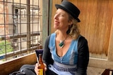 A smiling woman in a hat sitting on a bench drinking a beer.