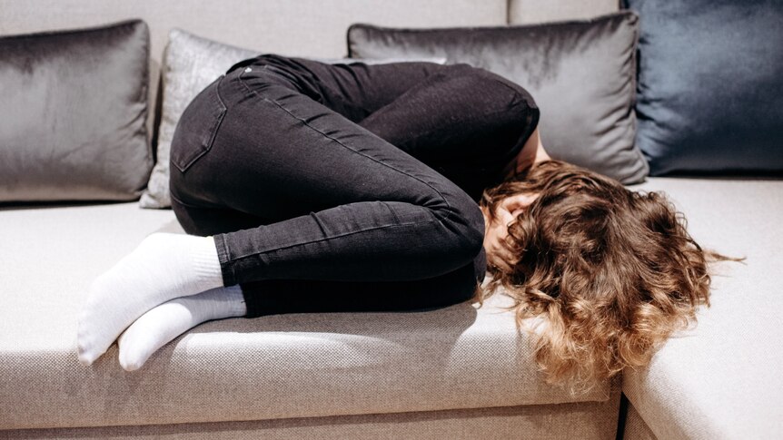 A child lays on a lounge, curled into the fetal position with their hands covering their face
