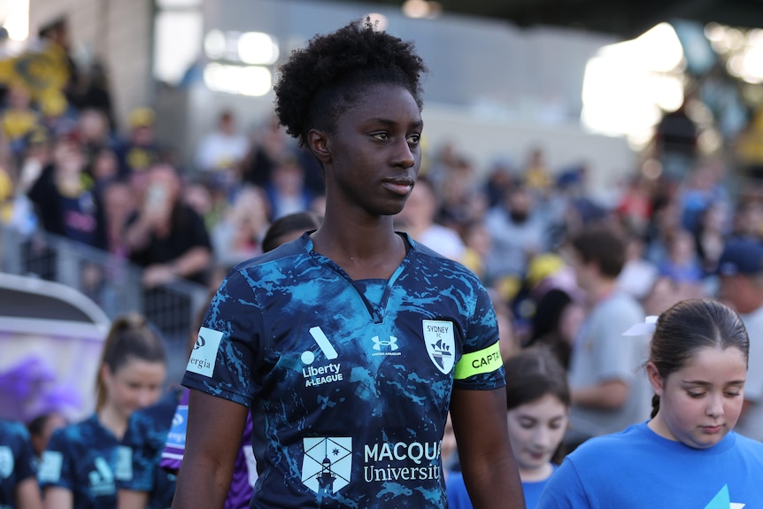 A soccer player wearing a dark blue marbled shirt walks out onto a field in front of a crowd