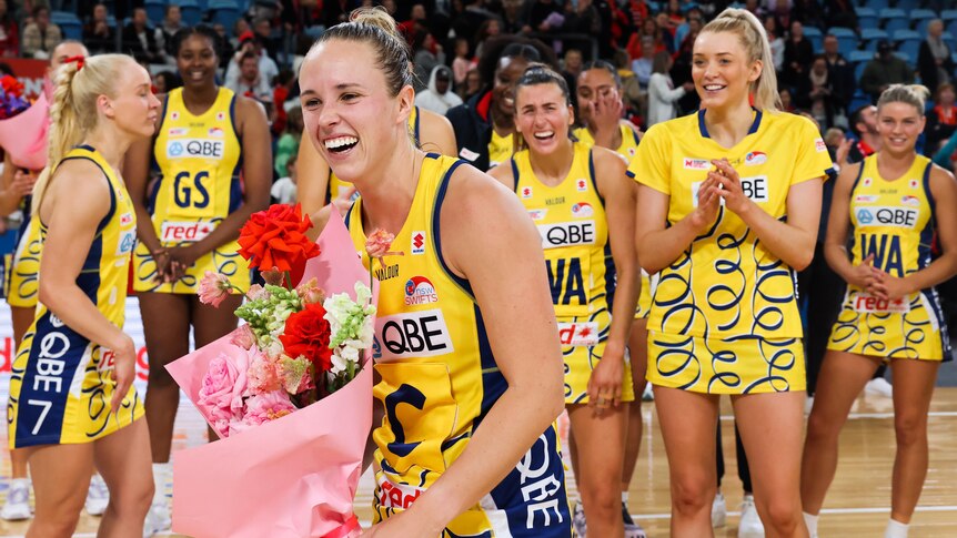 A Super Netball player smiles as she holds a bouquet of flowers, while her teammates cheer behind her. 