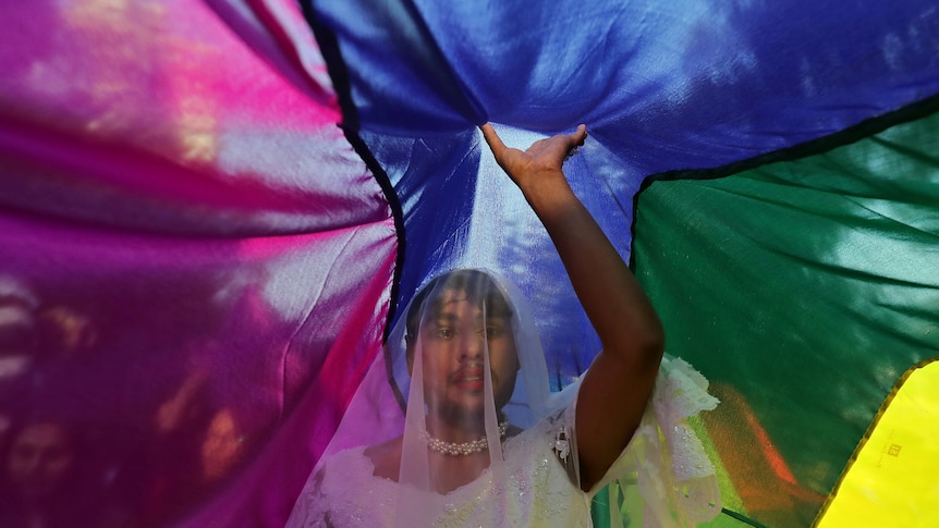 A person stand under a rainbow flag in a wedding dress