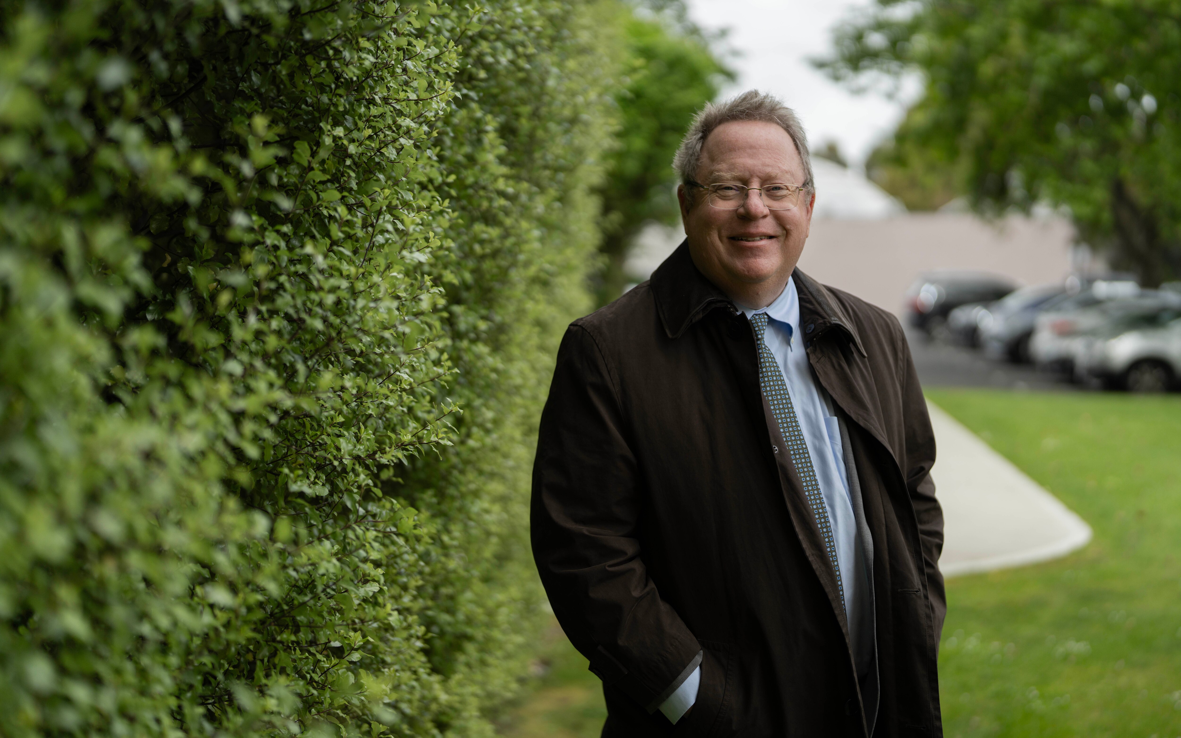 A man with glasses wearing a blue business shirt and brown coat.