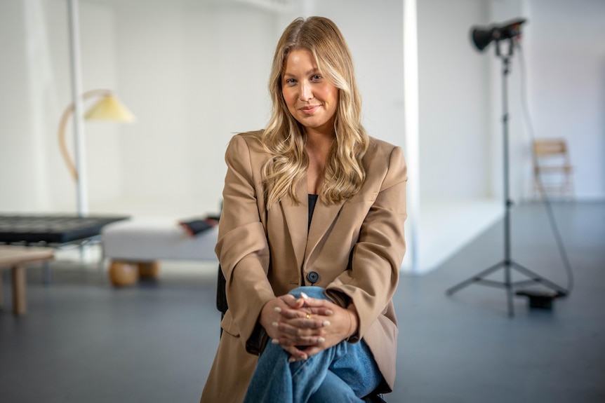 A woman sits with her legs crossed smiling at the camera with a photo shoot studio in the background