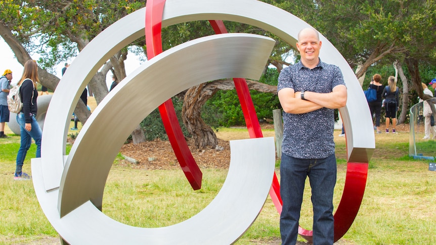 Balding man wearing short-sleeve shirt and jeans stands smiling with arms folded in front of sculpture made from steel loops.
