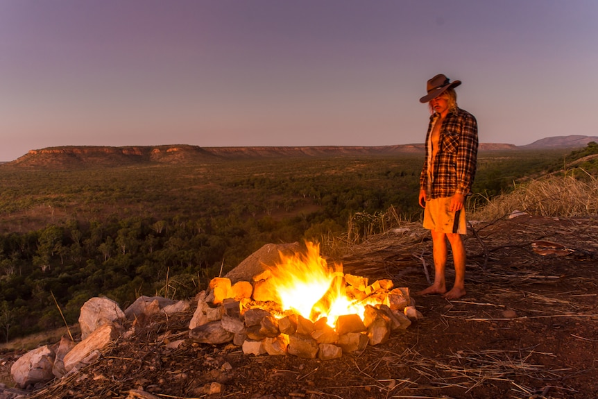 A man stands near a campfire with a flat-topped mountain range in the background