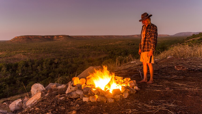 A man stands near a campfire with a flat-topped mountain range in the background