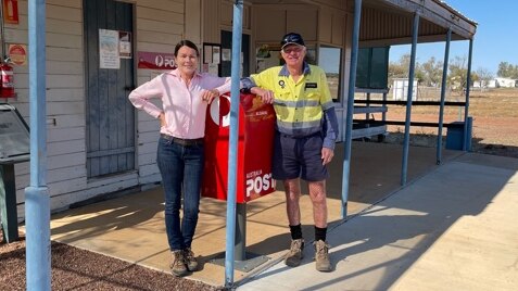 A man and a woman lean on a red post office box outside of a rural building.