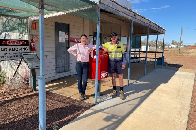 A man and a woman lean on a red post office box outside of a rural building.