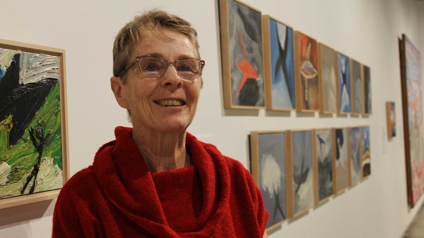 A woman in her 60's wearing glasses and a red top standing in an art gallery.