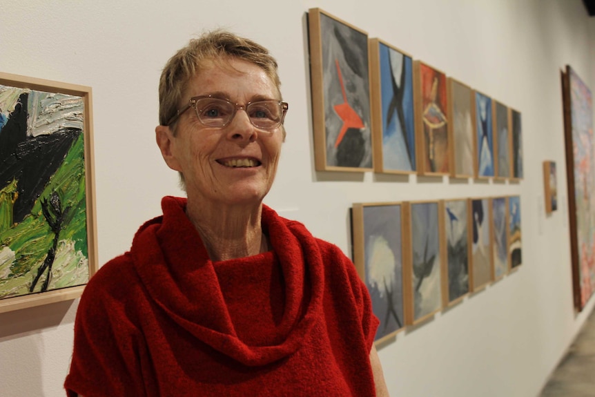 A woman in her 60's wearing glasses and a red top standing in an art gallery.