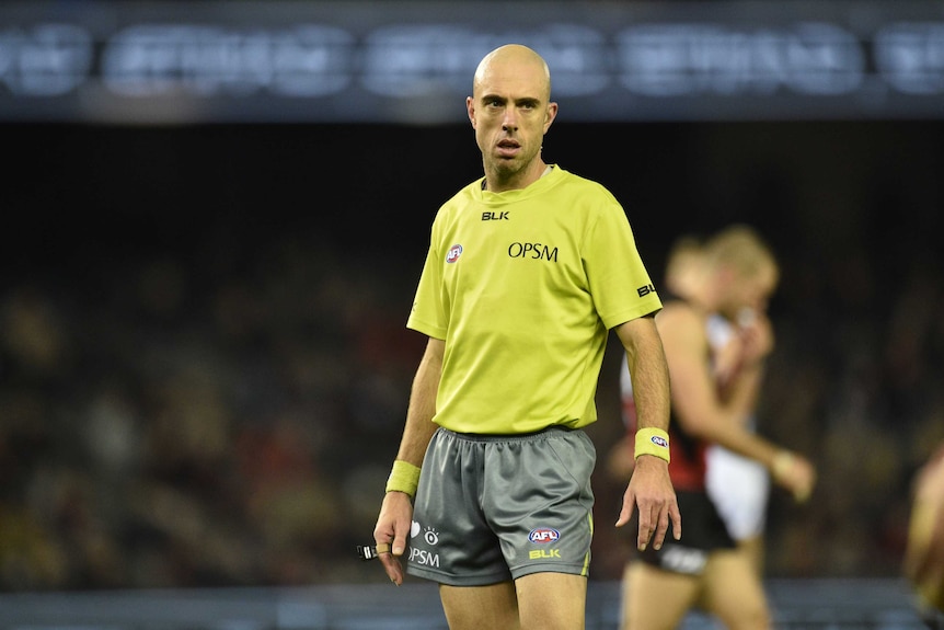 AFL umpire Mathew Nicholls stands and looks at the play while holding his whistle.
