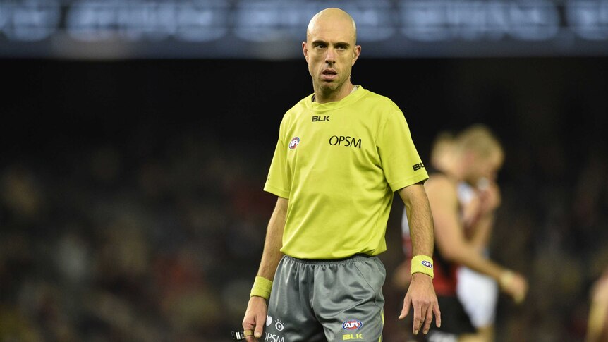 AFL umpire Mathew Nicholls stands and looks at the play while holding his whistle.