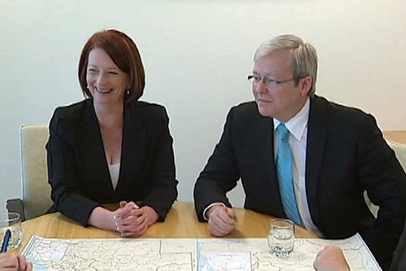 Julia Gillard smiles while Kevin Rudd looks forlorn, the two of them sitting side by side at a table looking at maps.