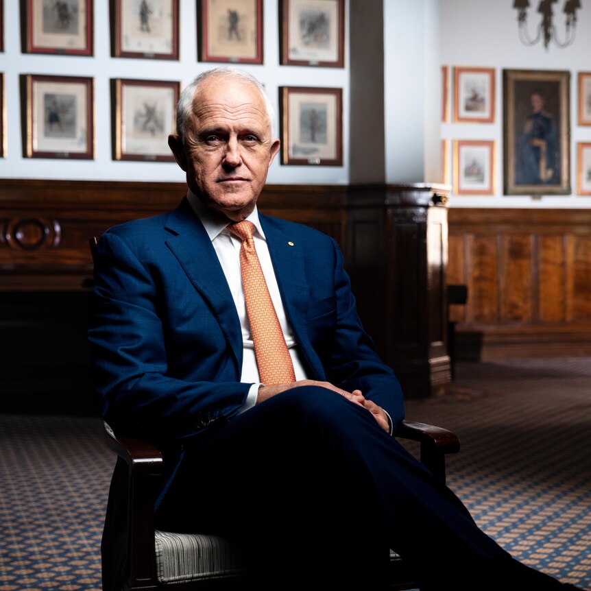 Dressed in a dark blue suit and orange tie, Malcolm Turnbull sits in a chair in a large wood-panelled room, looking serious.